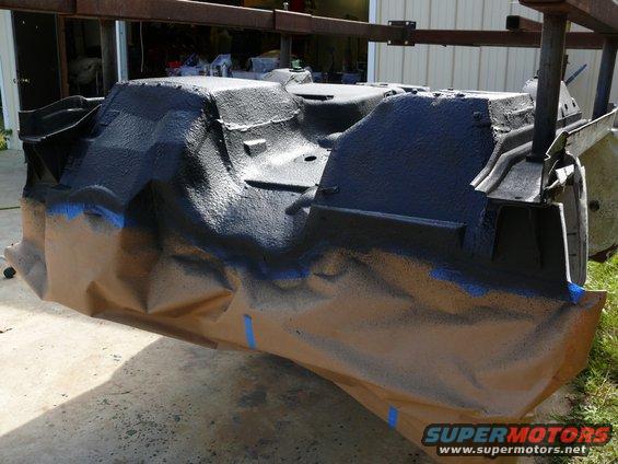 Car chassis part covered in black spray paint and masking paper, suspended by metal frame