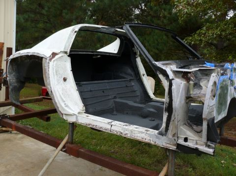 White 1975 Chevrolet Corvette body shell on a metal stand, side view, interior gutted, showing floor pan and d