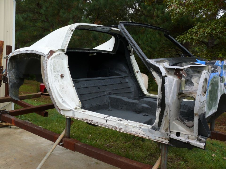 White 1975 Chevrolet Corvette body shell on a metal stand, side view, interior gutted, showing floor pan and door frame