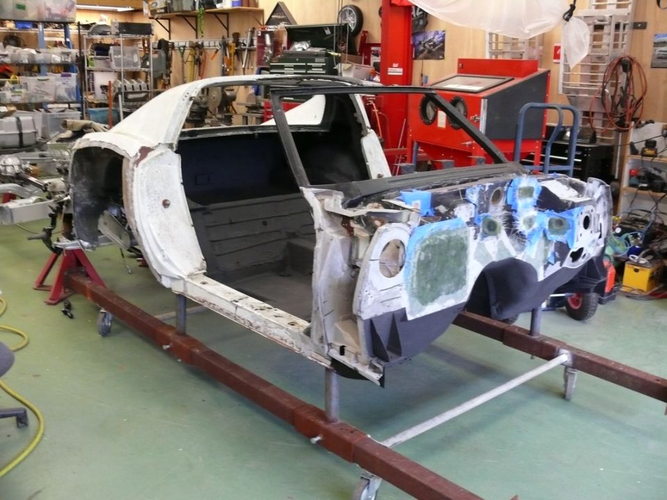 Car body on red stands in a workshop, undergoing repairs. White primer, exposed interior, and front end disassembled