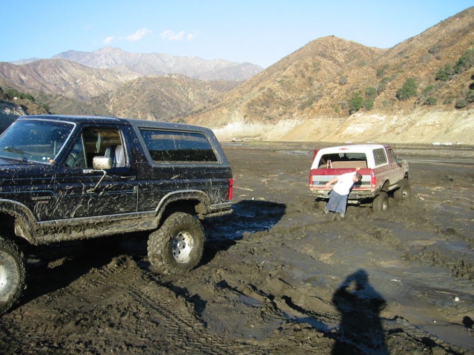 Muddy dark Ford Bronco on lifted tires in an off-road mud pit with a stuck red Bronco behind, mountains in background.
