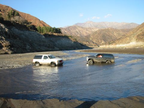 1985 Ford Bronco - Two SUVs navigate a riverbed, surrounded by arid, mountainous terrain under a clear blue sky.