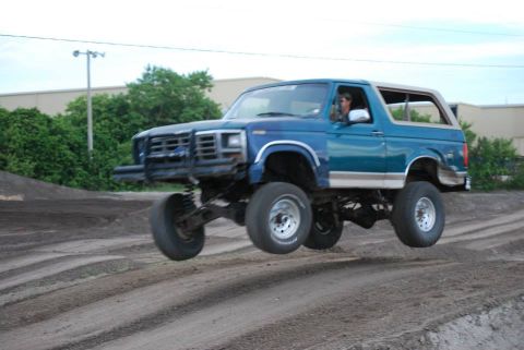1996 Ford Bronco - A lifted blue two-tone square-body Ford Bronco with a tan removable hardtop catches air over a dirt jump, all four wheels off the ground an…