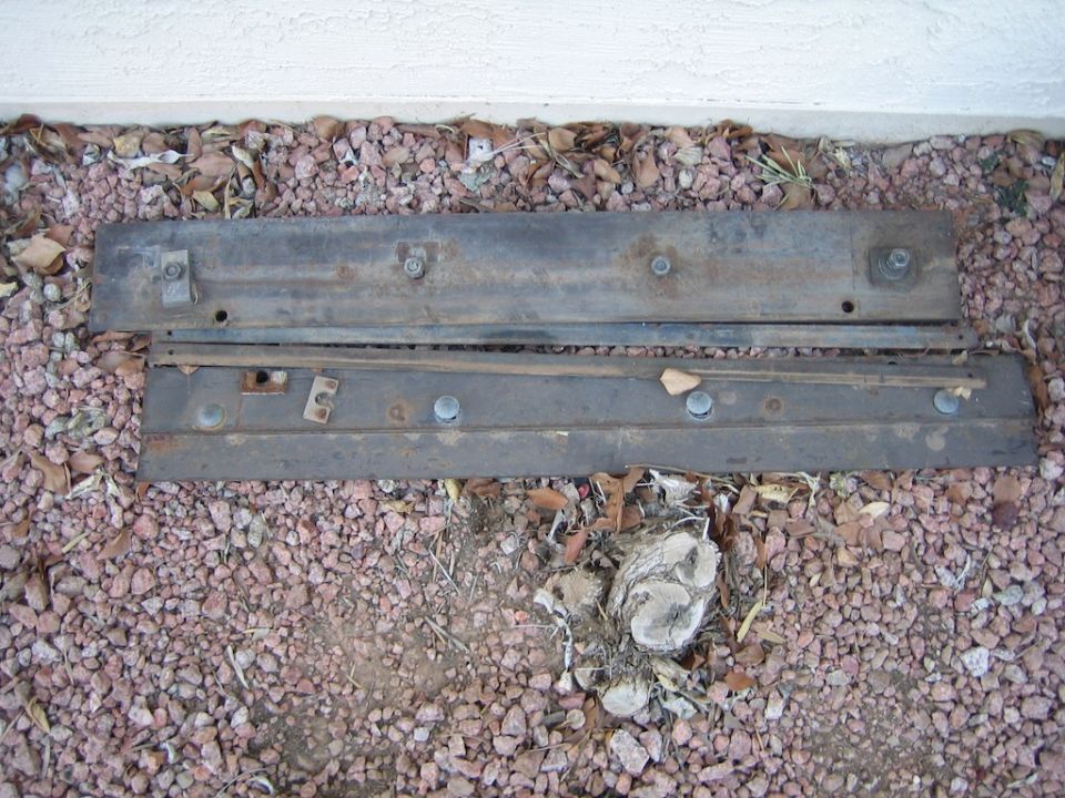 Two rusty metal car parts with bolts and brackets, lying on gravel and leaves