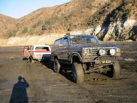 1985 Ford Bronco - Two off-road vehicles are stuck in thick mud on a dry lake bed with hills in the distance.