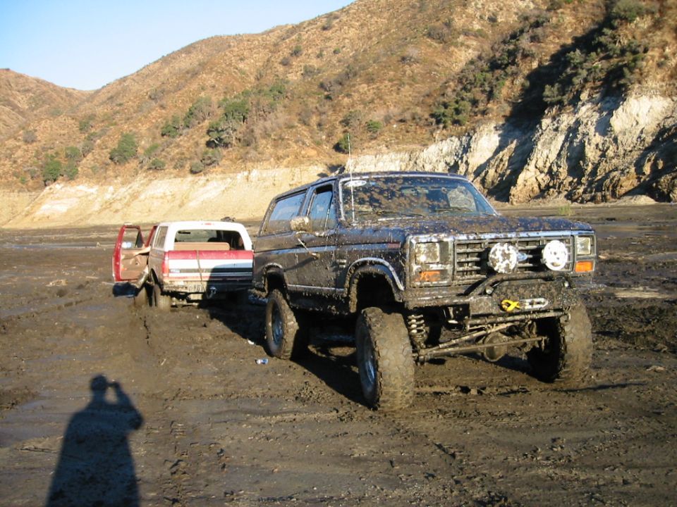 Muddy lifted dark Ford Bronco on big tires in a muddy riverbed with a stuck pickup behind it