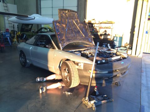 1995 Ford Probe - A mechanic works on a silver coupe with its hood and engine exposed, supported by a jack.