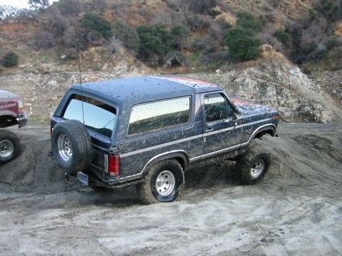 1985 Ford Bronco - A muddy Ford Bronco navigates a dirt incline, with another vehicle partially visible to the left.
