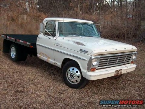 1994 Ford Bronco - White late-1960s Ford one-ton dually flatbed truck parked on leaf-covered ground in front of brush, seen from the front three-quarter with…