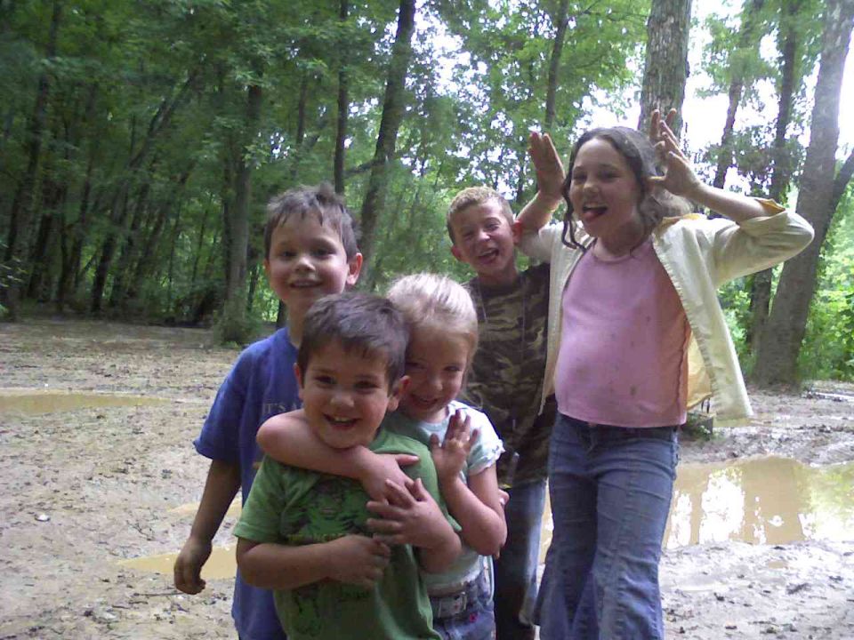 Five kids posing for a photo in a muddy, wooded area. Some are making funny faces