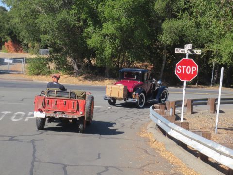 1946 Jeep CJ5 - Two vehicles, a red jeep-like vehicle and a maroon coupe, drive on a road near a stop sign.