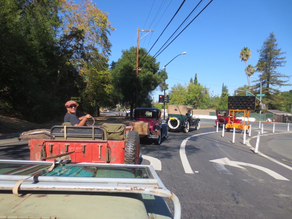 driver in flat cap in red jeep, other vintage cars behind on road