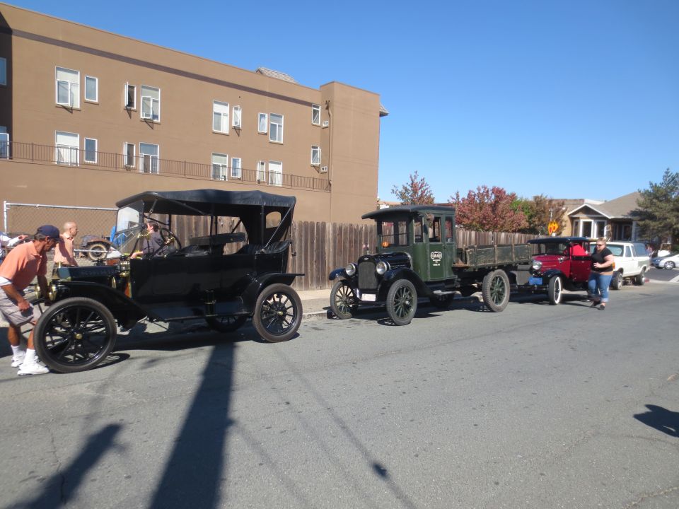 Three antique vehicles in a line on a street, a black touring car, a green truck, and a red truck