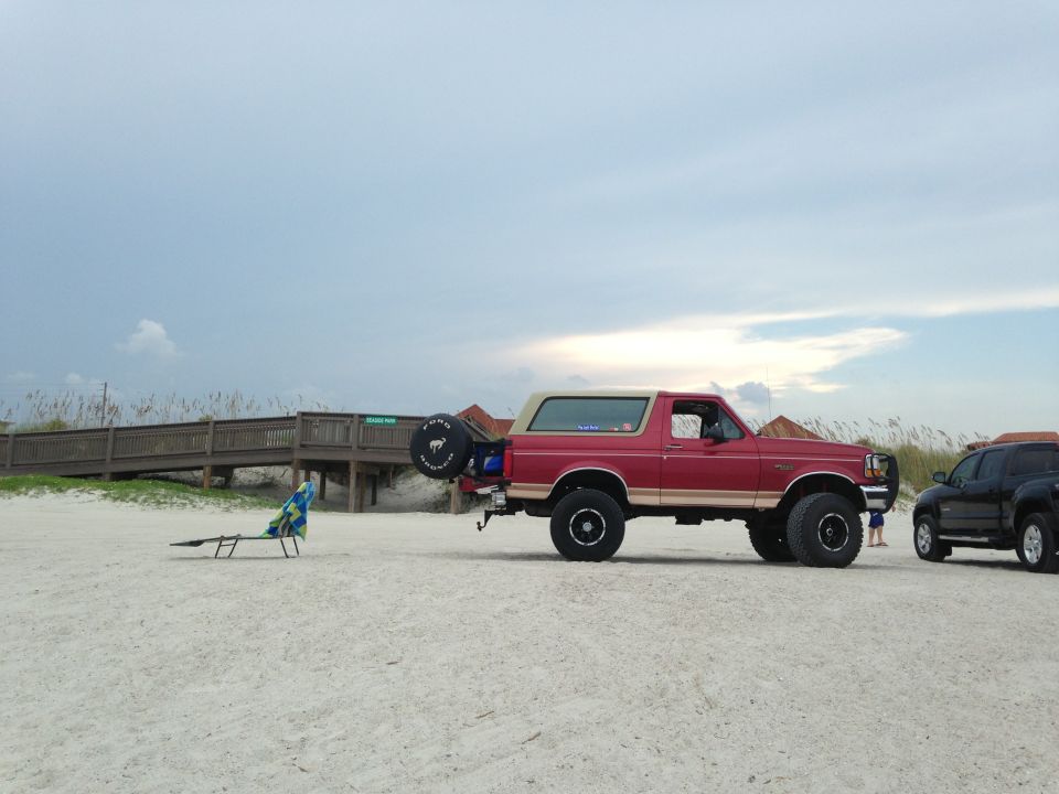 Red 1994 Ford Bronco parked on a sandy beach with a wooden boardwalk in the background. Another dark SUV is parked to the right