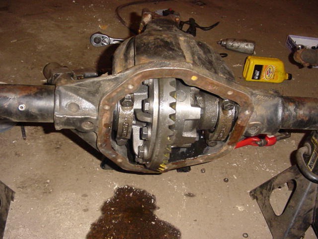 Differential axle assembly with open cover, gears visible, on concrete floor with tools and oil