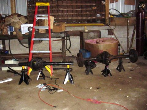 1992 Ford Bronco - Two vehicle rear axles are supported by jack stands in a cluttered workshop setting, with a ladder and various tools in the background.