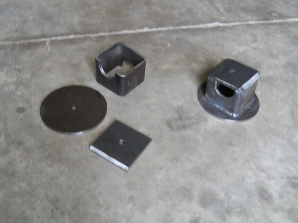Four metal parts on concrete floor: a circular plate, a square plate, and two square brackets with openings