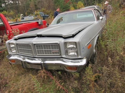 1991 Ford LTD Crown Victoria - A silver sedan, likely a 1977 model, sits in a junkyard with its hood ajar, surrounded by overgrown vegetation and other vehicles.