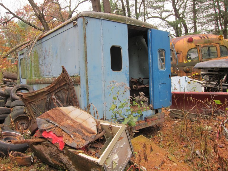 Abandoned blue step van with open doors, next to a yellow school bus and other scrap metal in a junkyard