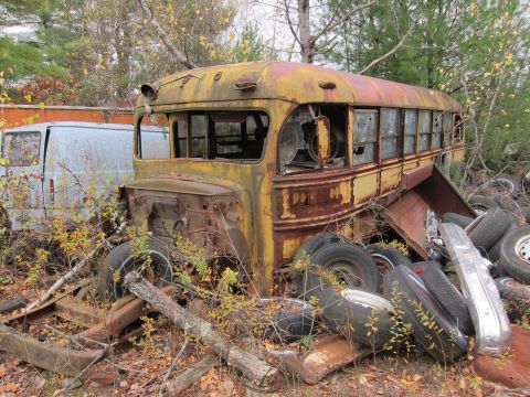 1991 Ford LTD Crown Victoria - An abandoned school bus sits in a junkyard, surrounded by debris and other vehicles.