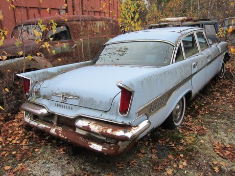 1991 Ford LTD Crown Victoria - A derelict 1957 Chrysler New Yorker sedan sits in a junkyard, surrounded by fallen leaves and other decaying vehicles.