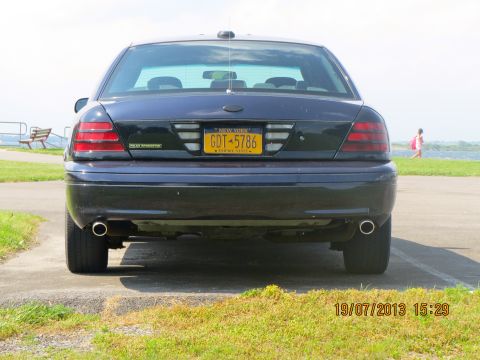 2003 Ford Crown Victoria - A dark sedan is parked outdoors near a body of water, with a person walking in the background.