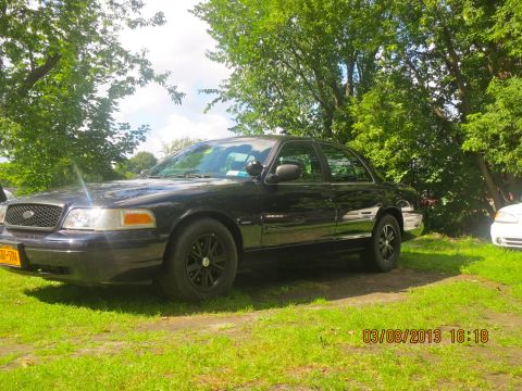 2003 Ford Crown Victoria - A black 2003 Ford Crown Victoria is parked on a grassy area with trees in the background. Another vehicle is partially visible to the right.