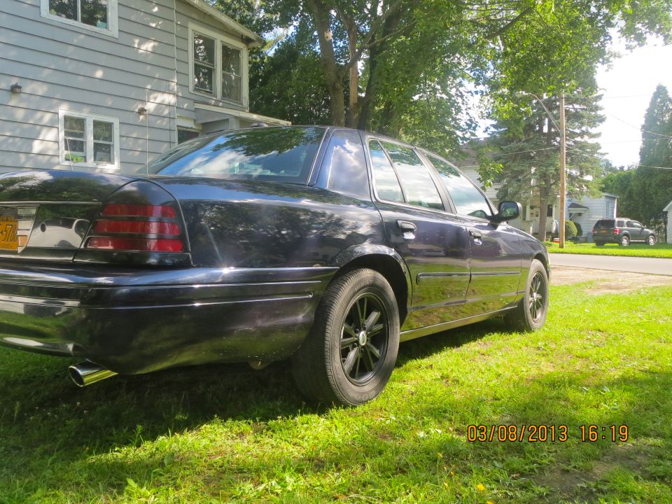 Dark sedan parked on grass in front of a house. Another SUV is visible in the background