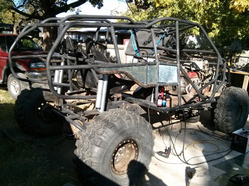 Custom off-road buggy frame with roll cage and large tires, red truck in background