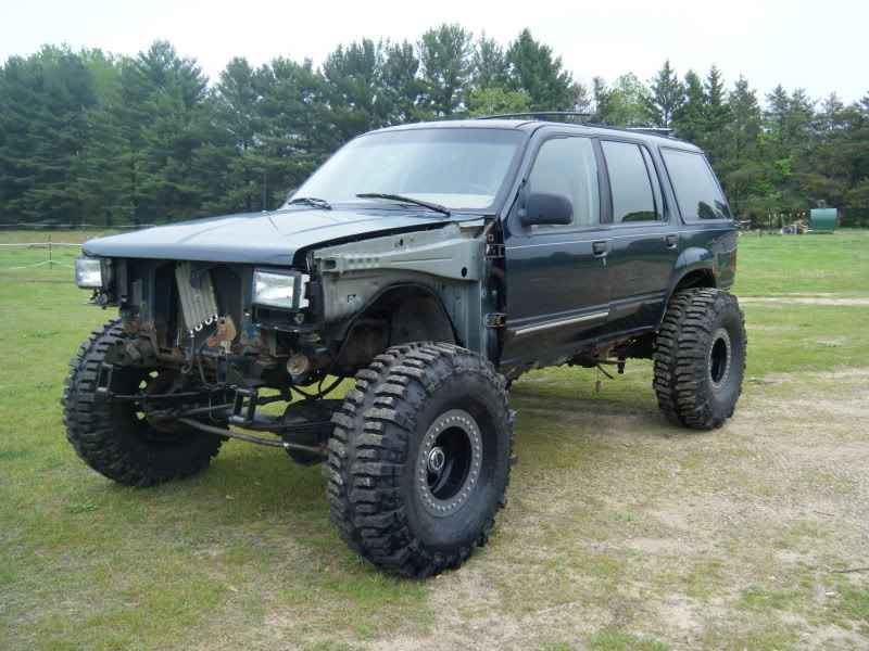 Modified dark green SUV with large off-road tires, no front fenders, parked on grass.