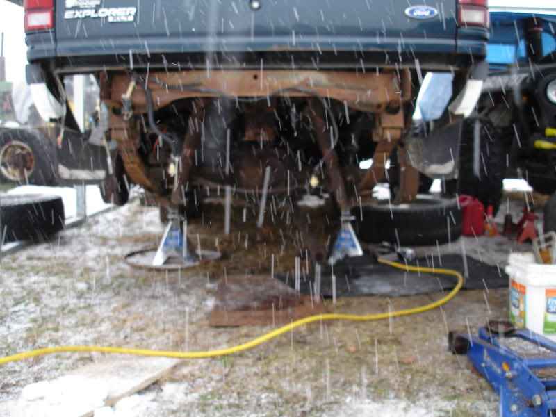 Rear view of a dark blue Ford Explorer on jack stands in the snow, with yellow hose on the ground.
