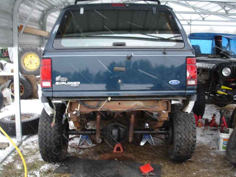 Rear view of a dark blue Ford Explorer SUV on jack stands in a garage during snowfall.