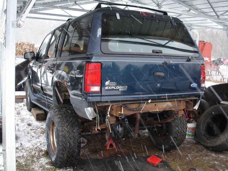 Rear driver's side of a lifted dark blue Ford Explorer with large tires, missing rear bumper, in the snow.