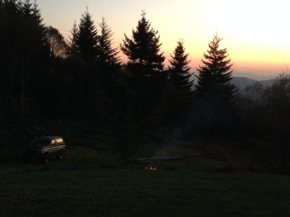 1989 Ford Bronco parked in a clearing with a campfire at dusk.