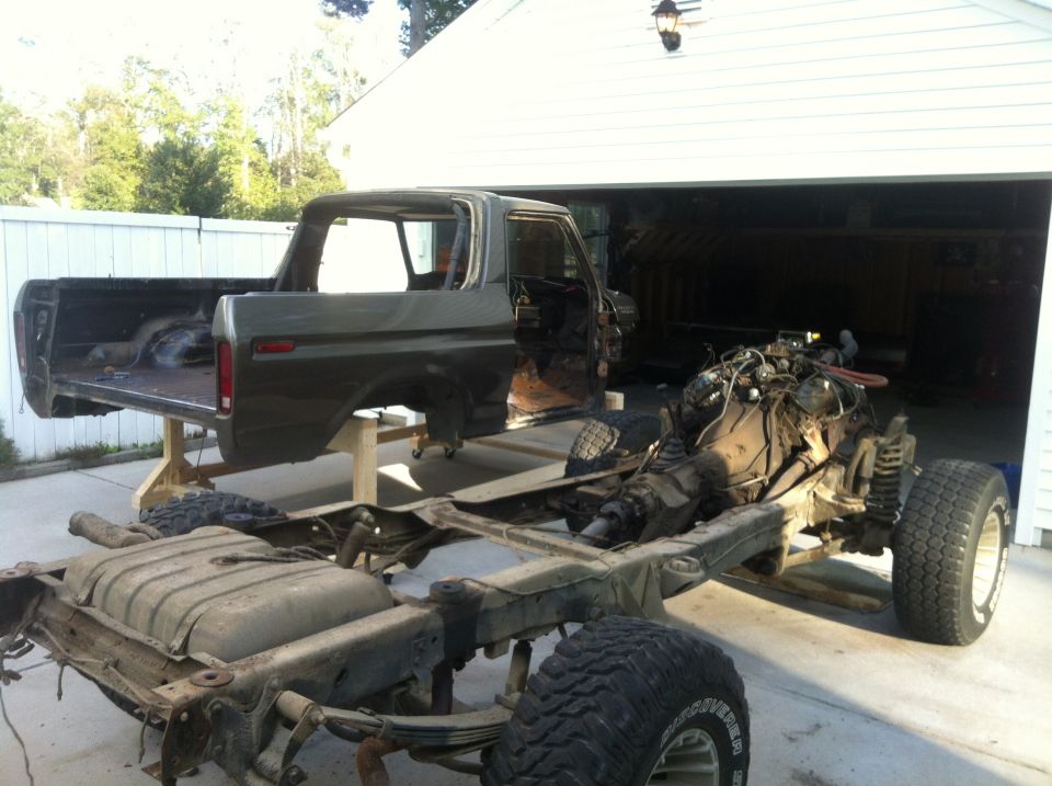 1979 Ford Bronco chassis and body disassembled outdoors in front of a garage.