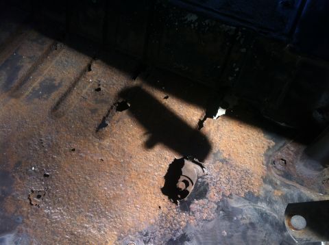 1979 Ford Bronco - The underside of a vehicle shows significant rust and holes in the floor pan. A shadow of an object is cast across the corroded metal.