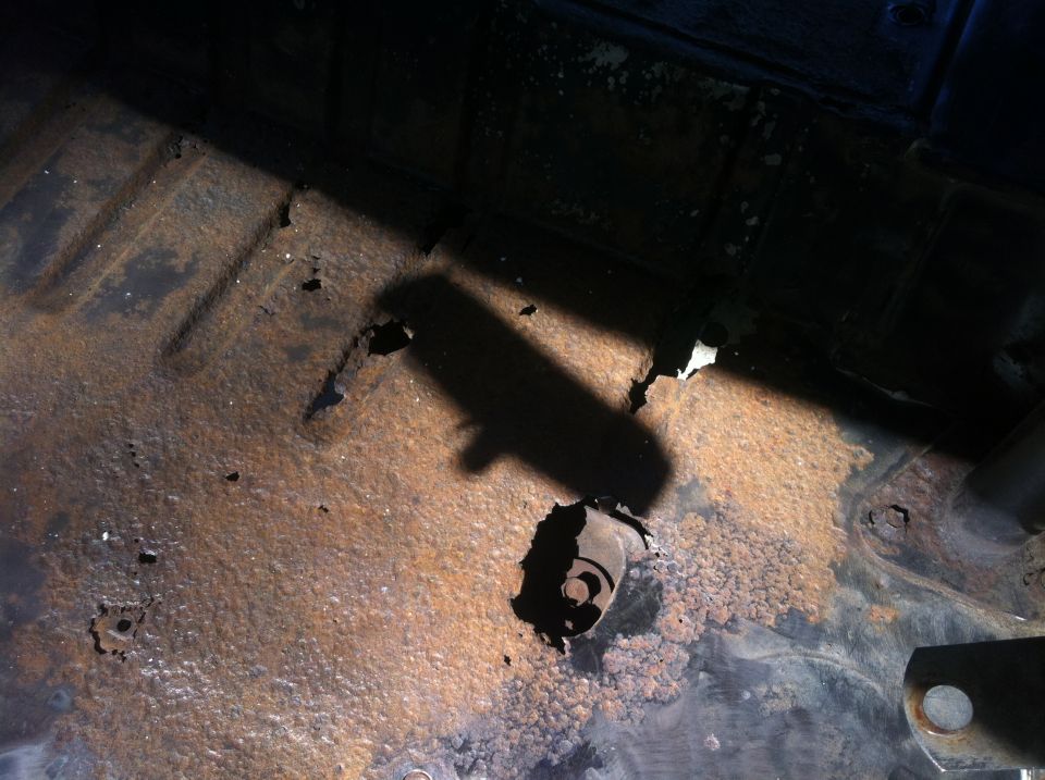 Underside of vehicle with severe rust and holes in floor pan, shadow cast across.