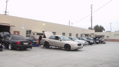 2003 Ford Crown Victoria - Multiple full-size sedans, hoods open, line up along a tan industrial building while people tend to them in an outdoor lot.