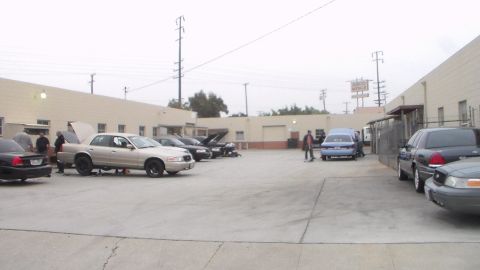 2003 Ford Crown Victoria - wide overcast view of a concrete industrial lot with several sedans parked along tan buildings and a metals-yard sign in the distance.