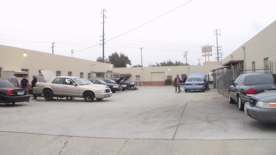 Wide view of a paved industrial lot with sedans parked along buildings under an overcast sky