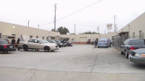 2003 Ford Crown Victoria - concrete lot framed by tan warehouses holds several sedans, a few with hoods raised, with a blue car parked across the yard.