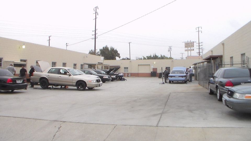 Industrial parking lot with multiple sedans, some with hoods open, beside tan warehouse buildings