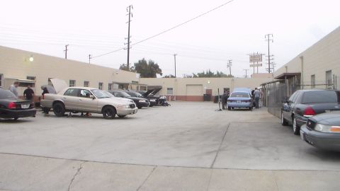 2003 Ford Crown Victoria - Several full-size sedans with hoods up sit in a wide concrete lot near tan buildings while people gather around a blue car at right.