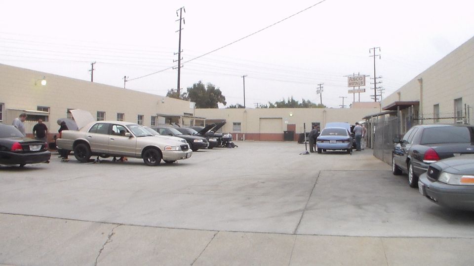 Sedans with raised hoods parked in a wide concrete industrial lot under gray sky