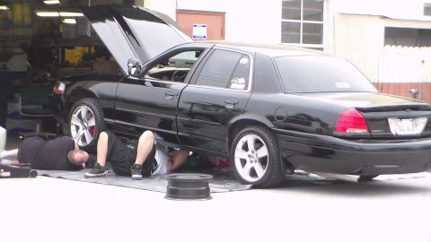 2003 Ford Crown Victoria - black full-size sedan sits with its hood raised as two people lie on the ground working underneath, a bare wheel rim on the pavement nearby.