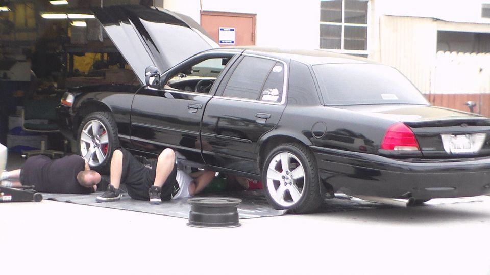 Black full-size sedan with hood up and two people lying underneath working on it
