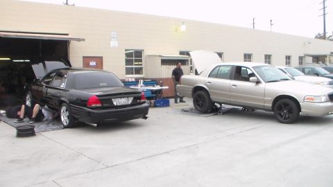 2003 Ford Crown Victoria - black sedan partly inside a shop bay sits beside a tan full-size sedan with its hood raised, with a person lying under the black car.