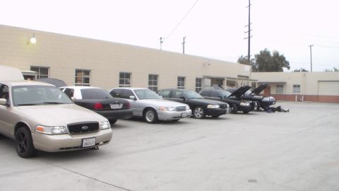 2003 Ford Crown Victoria - row of full-size sedans parked against a tan building; the nearest tan car shows a Ford oval on its grille.