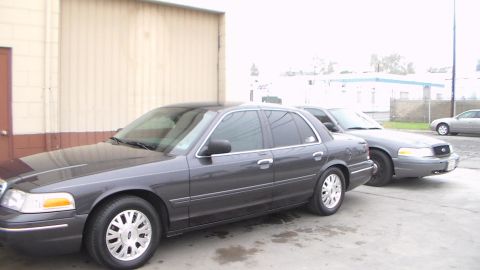 2003 Ford Crown Victoria - dark gray full-size sedan with chrome-style wheels is parked at an angle beside a tan sedan along a tan building.