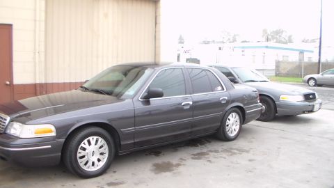 2003 Ford Crown Victoria - dark gray full-size sedan sits next to a tan car showing a Ford oval badge, parked along a tan building under gray sky.